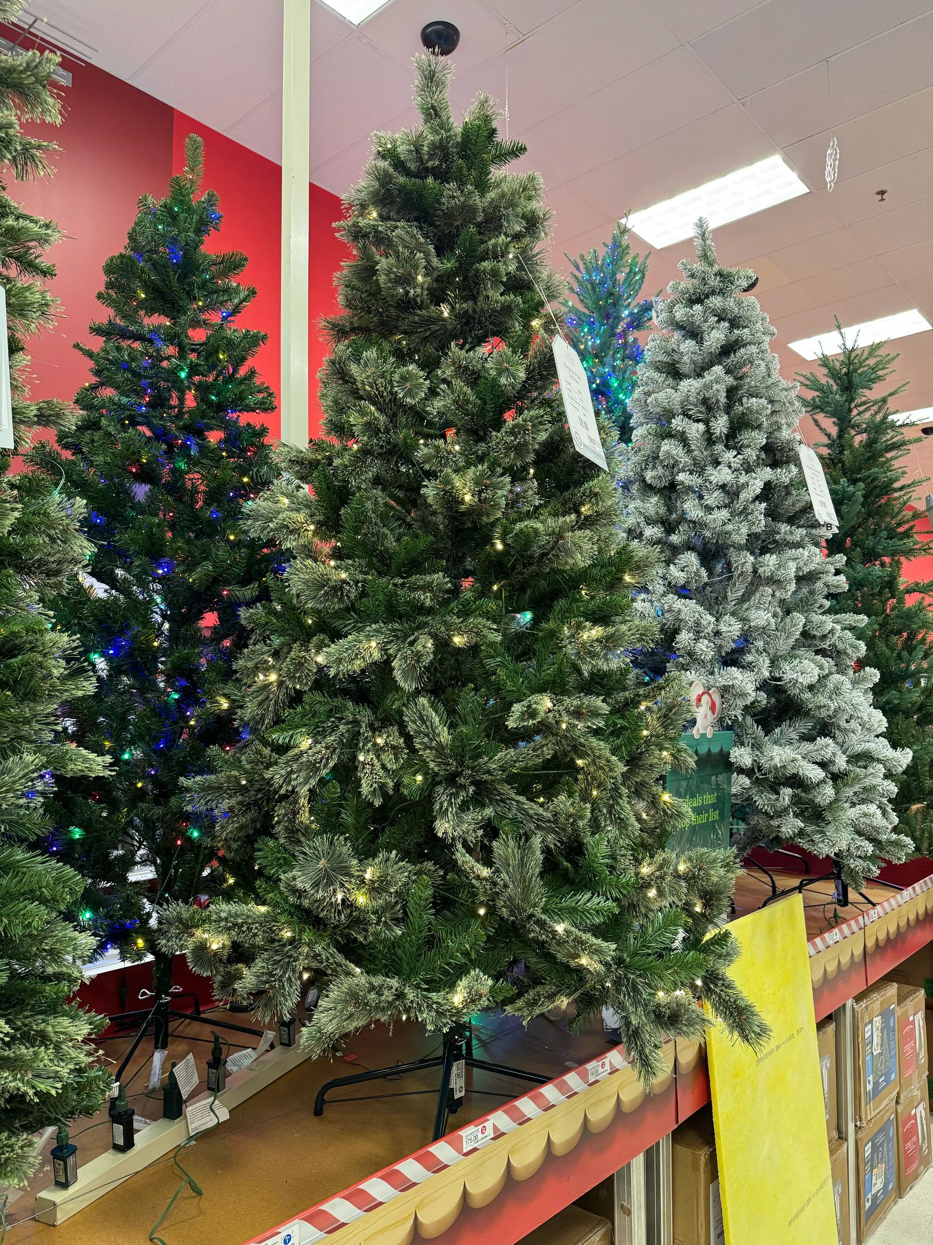 Pine-style trees with traditional PVC needles, moderate branching density, multicolor mini bulbs and warm LEDs, visible gaps between branches, budget construction quality