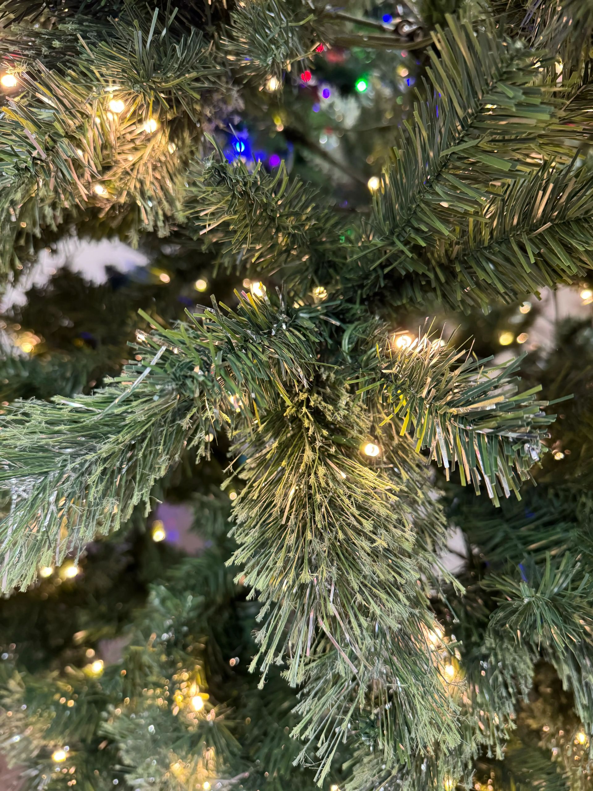 PVC pine branches with silver tinsel strands throughout, warm white mini bulbs, multicolor lights visible in background