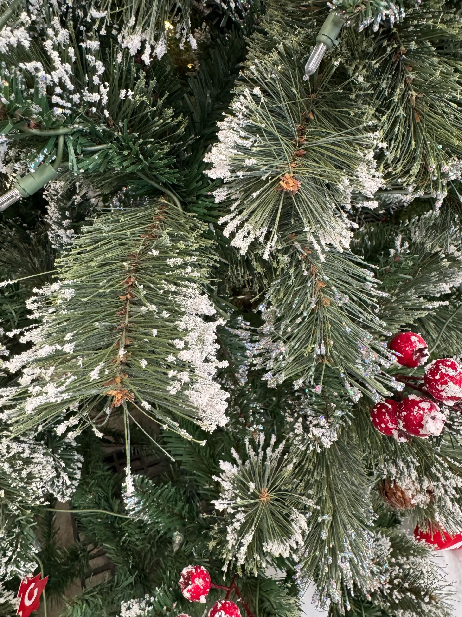 Dense pine needles heavily dusted with white glitter snow, red frosted berries, warm white LED lights visible between branches