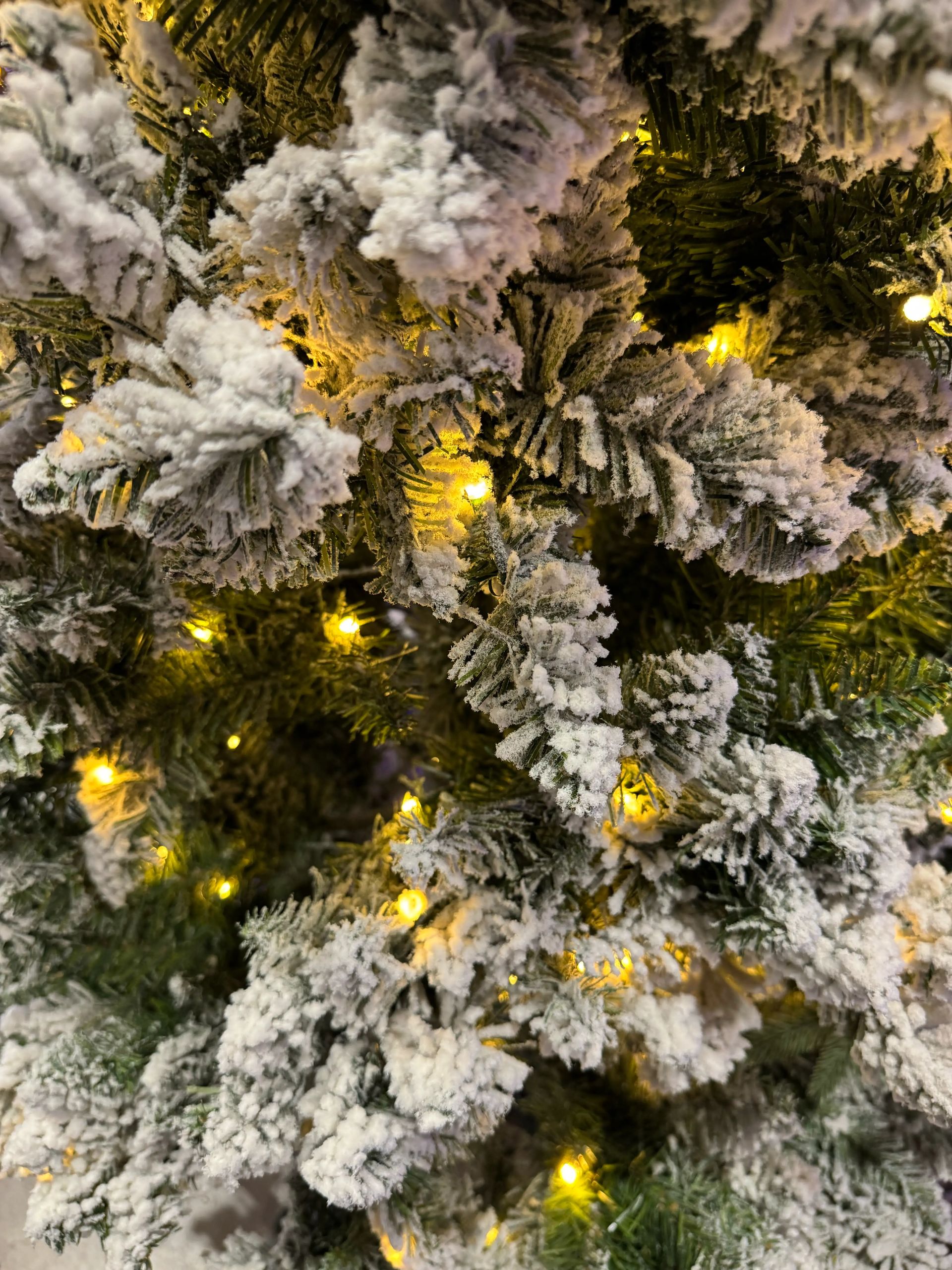 Heavy white flocking on PVC spruce branches, warm yellow micro LEDs glowing throughout, dense snow coverage on tips