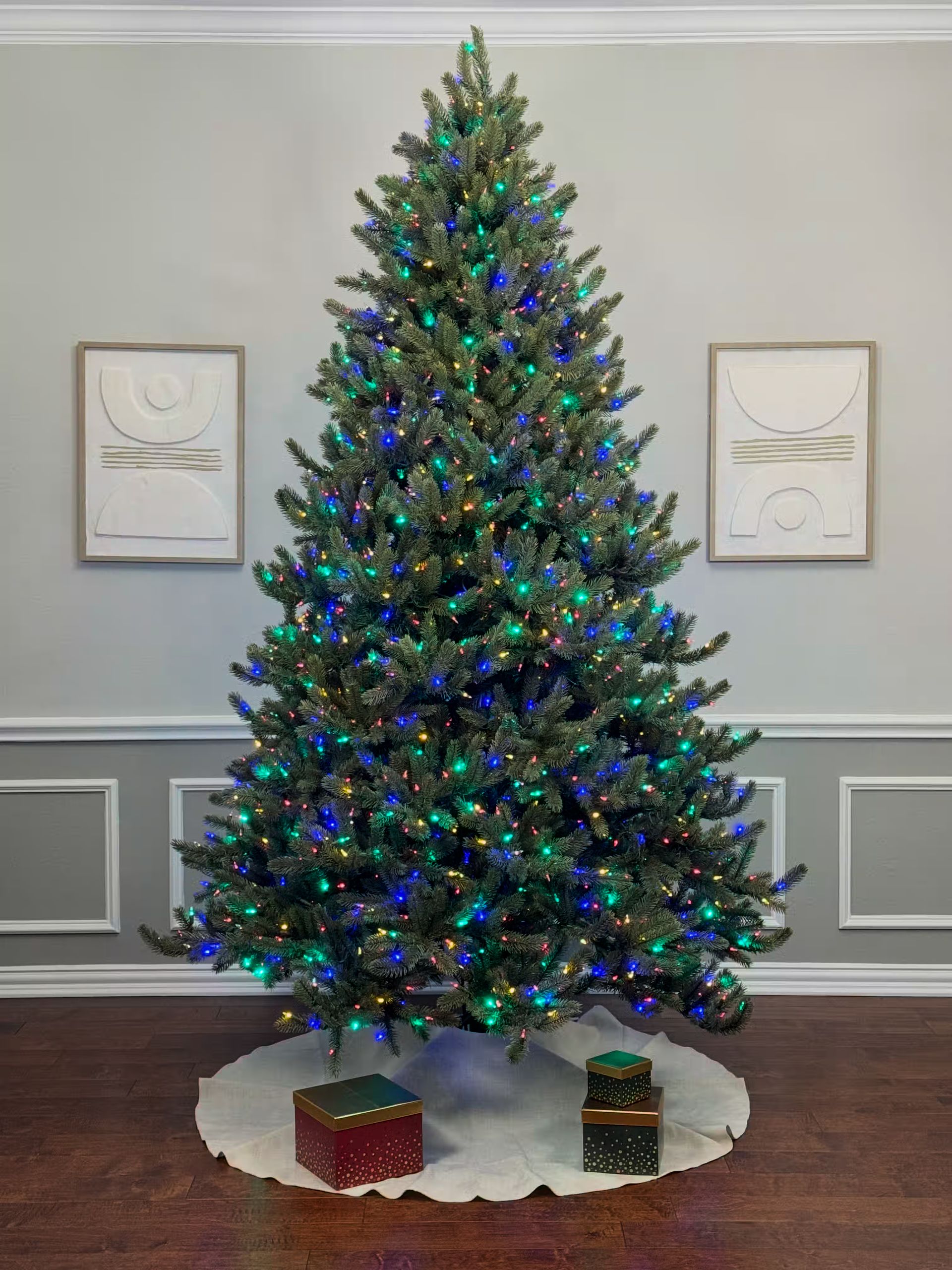 Room lights on with a very dense 8ft tree with color lights. The lights are so vibrant they create a cool blue cast on the wall.