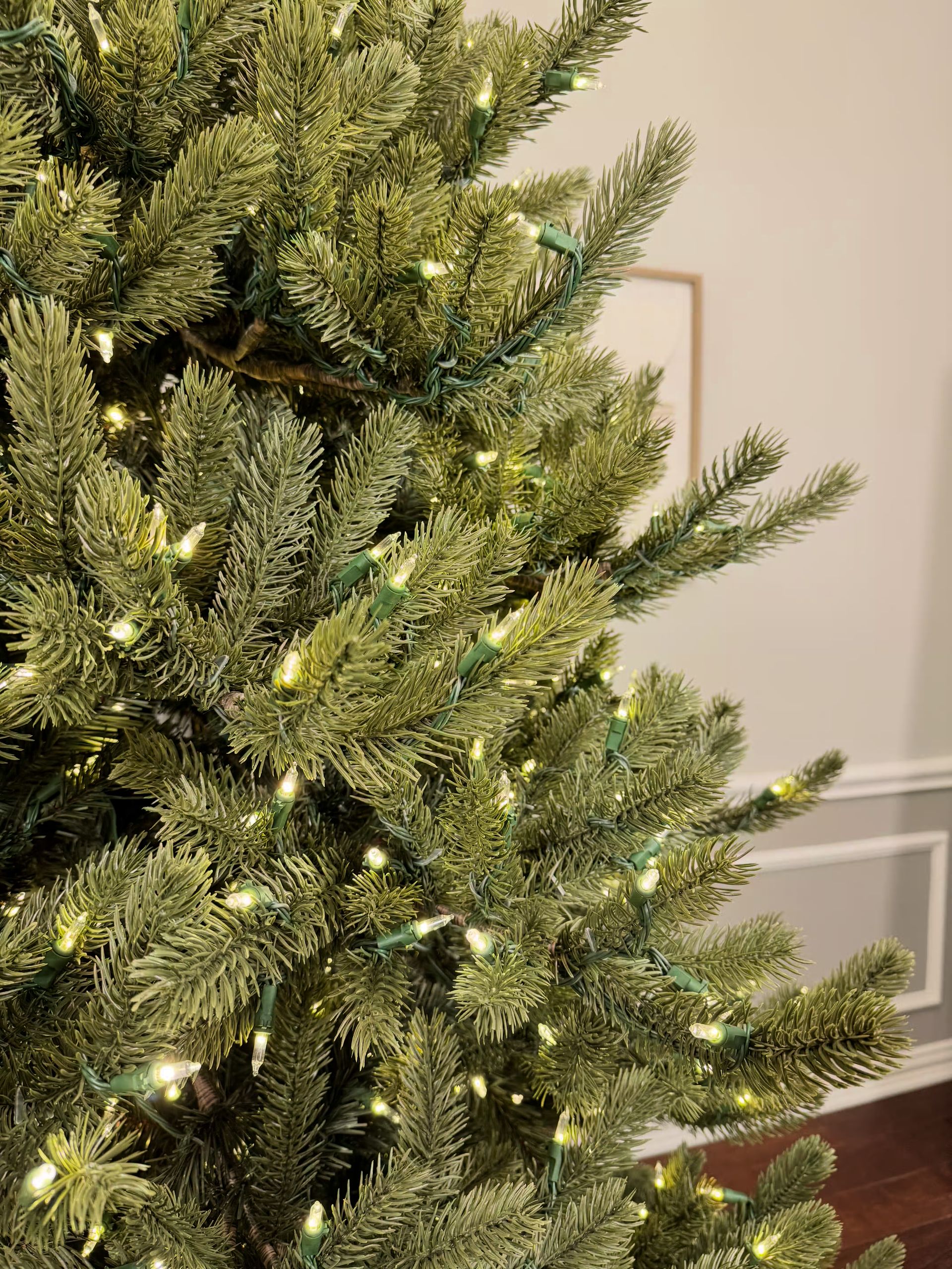 Room lights on with a close up of branch tips with clear lights and the wall in the background.